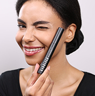 Une femme sourit vivement tout en tenant une brosse à mascara, démontrant son enthousiasme pour la beauté et l'application du maquillage.