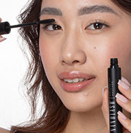 On voit une femme appliquer du mascara sur ses yeux, tenant délicatement la brosse tout en rehaussant ses cils.