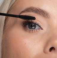 Une femme applique soigneusement du mascara sur ses cils à l’aide d’une brosse à mascara, rehaussant ainsi son maquillage des yeux avec précision.